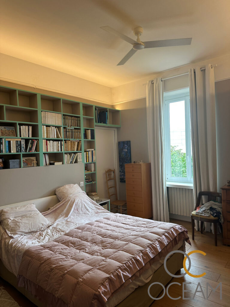 Ventilateur de plafond blanc à 3 pales, suspendu dans une chambre meublée d'un lit, d'étagères de livres et d'une fenêtre aux rideaux gris.