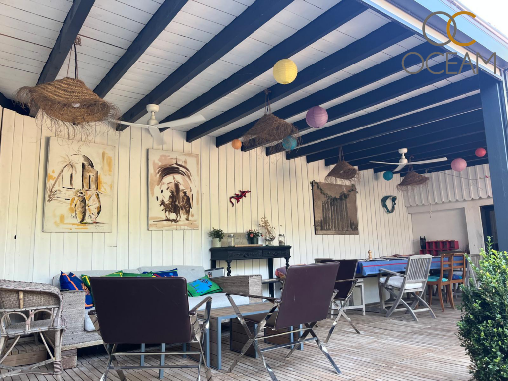 Ventilateur de plafond blanc à 5 pales sous un toit en bois noir et blanc, dans une terrasse de restaurant avec lampes en paille et lanternes colorées.