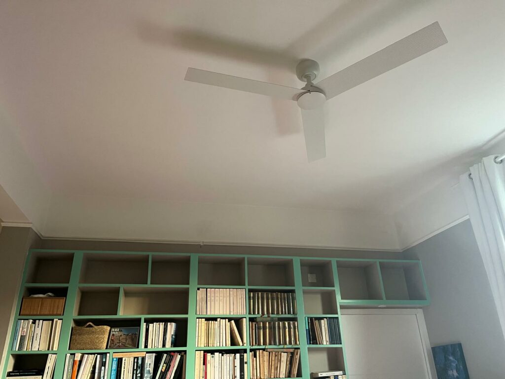 Ventilateur de plafond blanc à 3 pales fines, installé au-dessus d’une bibliothèque murale verte remplie de livres.