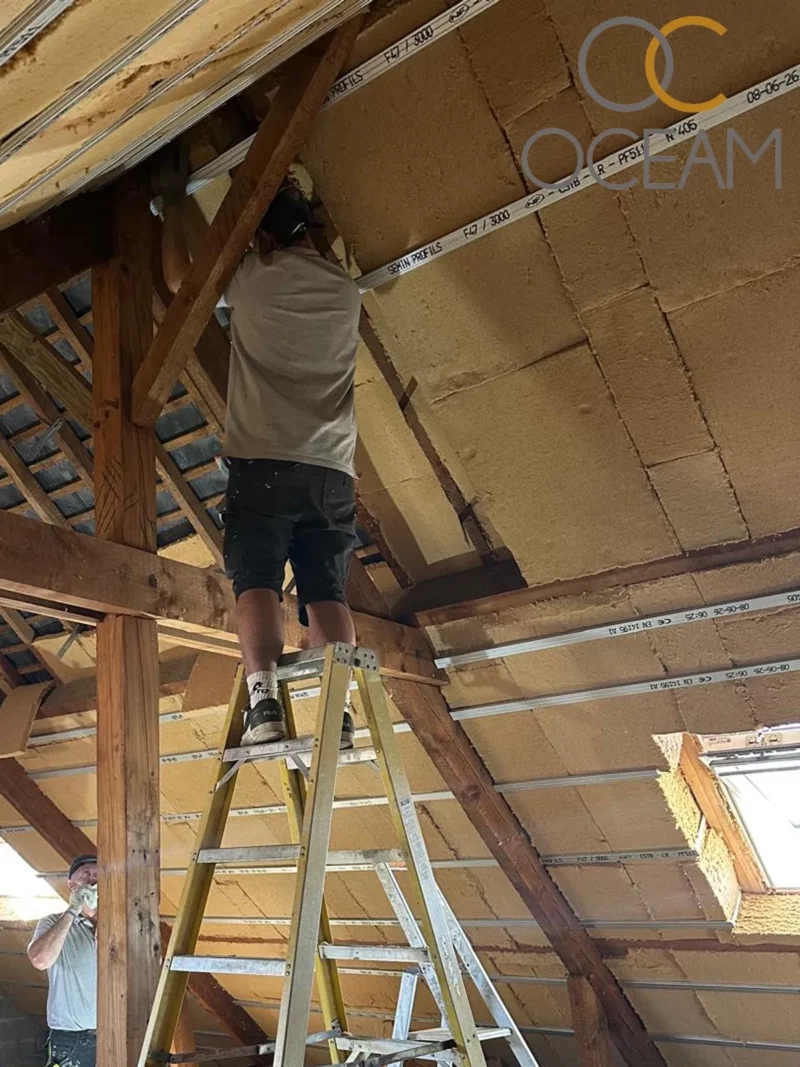Isolation sous rampants de toiture à La Chapelle-des-Marais — chantier OCEAM. Compagnon d'OCEAM sur échelle fixant des panneaux de fibre de bois entre les poutres du toit à La Chapelle-des-Marais